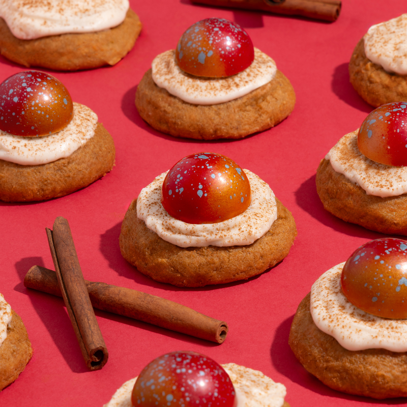 A close-up of Pumpkin Spice Cookie chocolates sitting on frosted cookies on a red background. Two crossed cinnamon sticks sit among the cookies.